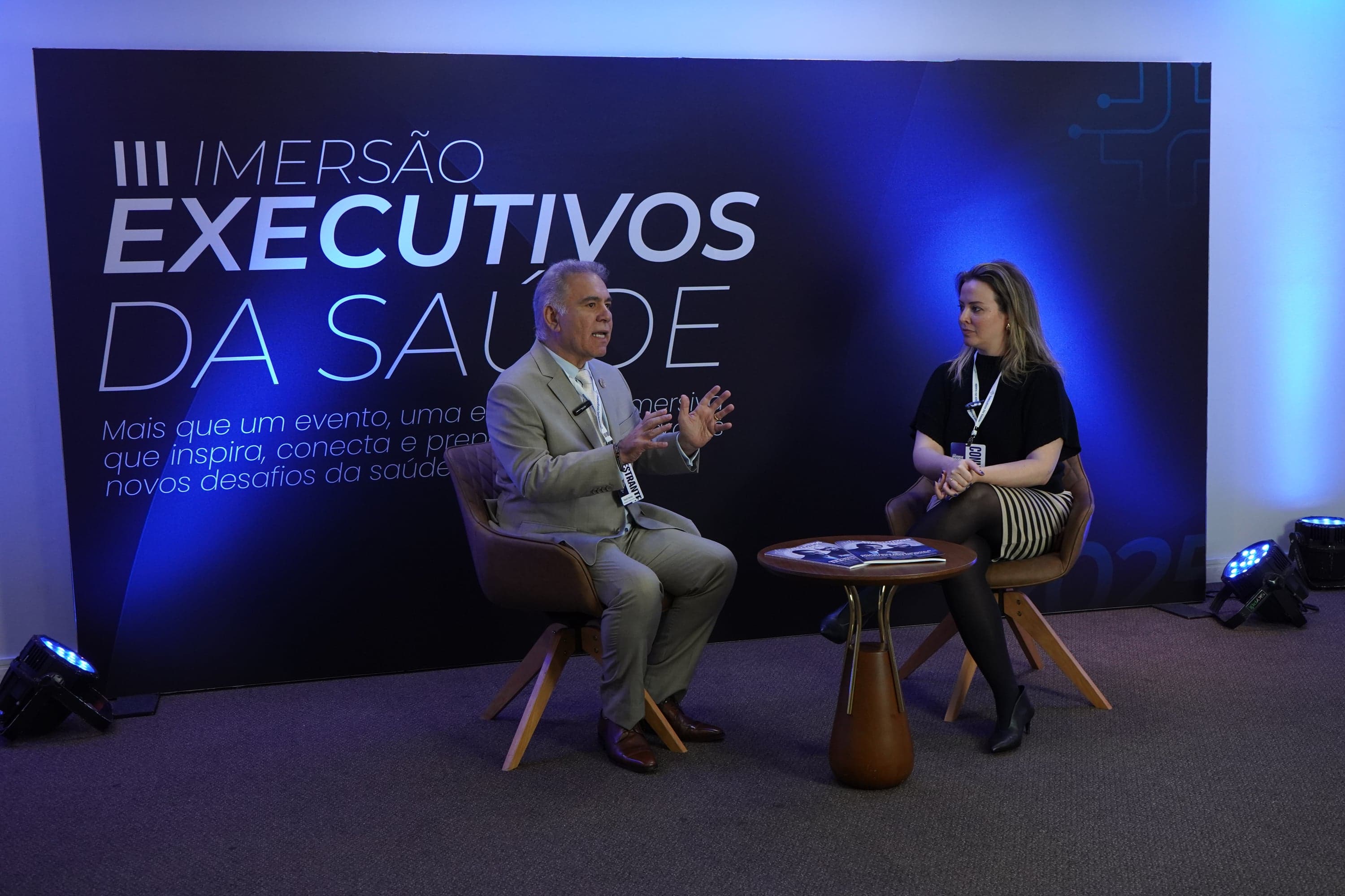 Dois entrevistados conversam sentados em poltronas no palco, diante do painel oficial 'III Imersão Executivos da Saúde', em formato de entrevista gravada