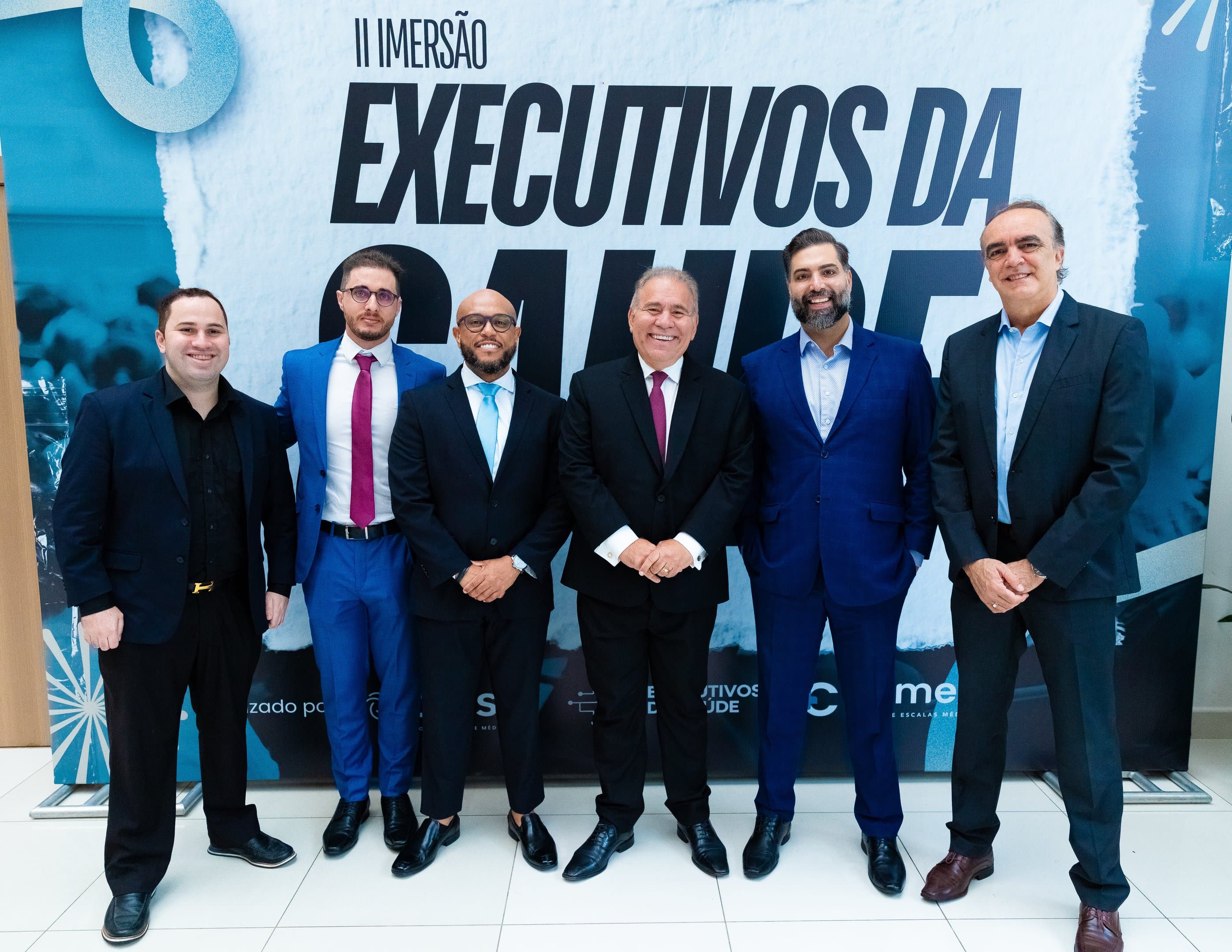 Grupo de executivos e organizadores posando diante do painel oficial de uma edição anterior da Imersão Executivos da Saúde